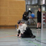 Ein Schüler wirft den Goalball und seine Mitspielerin befindet sich in der Verteidigungsposition. Beide tragen Dunkelbrillen.