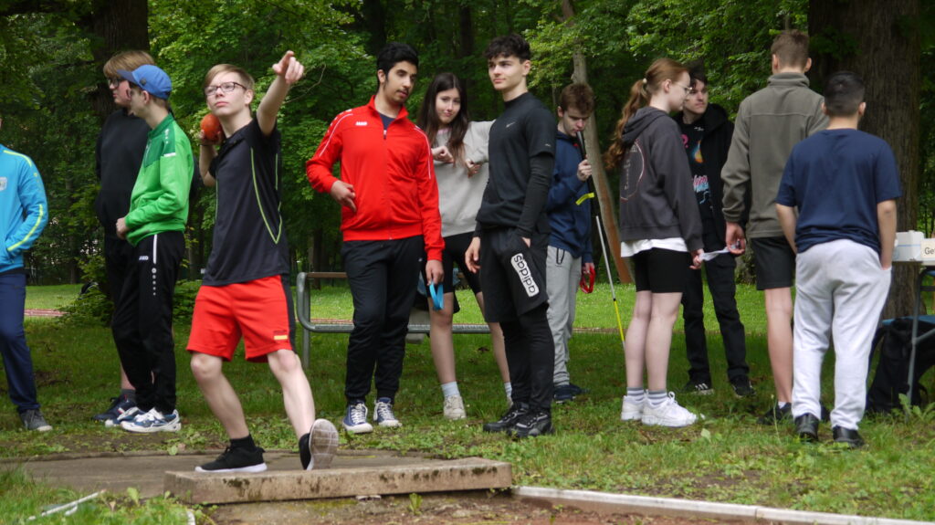 Ein Schüler steht beim Kugelstoßen bereit und hält die Kugel bereits in der Ausgangsstellung fest. Im Hintergrund warten andere Schüler auf ihren Einsatz.