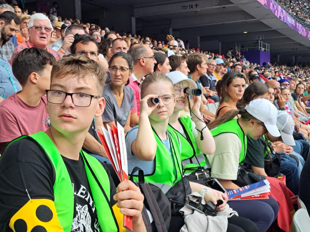 Zwei Schülerinnen im Stadion nutzen ihr Monokular, um das Wettkampfgeschehen zu verfolgen.