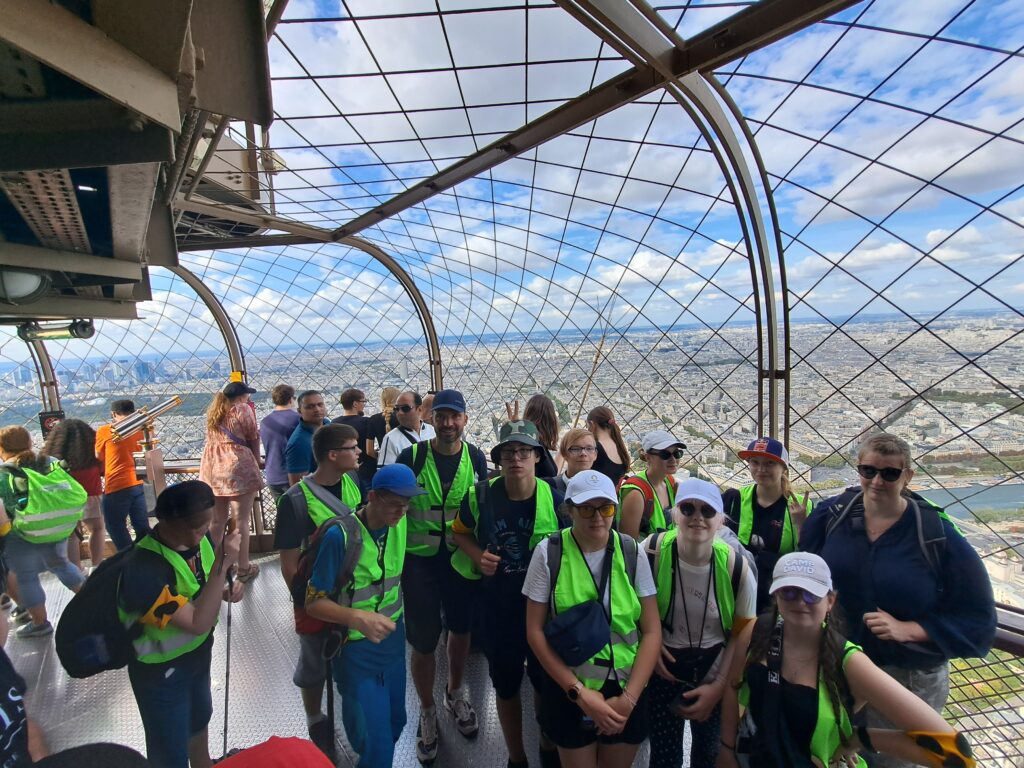 Gruppenfoto auf der Spitze des Eiffelturms. Im Hintergrund ist ein Großteil der Stadt Paris zu sehen. Die Häuser wirken sehr klein.