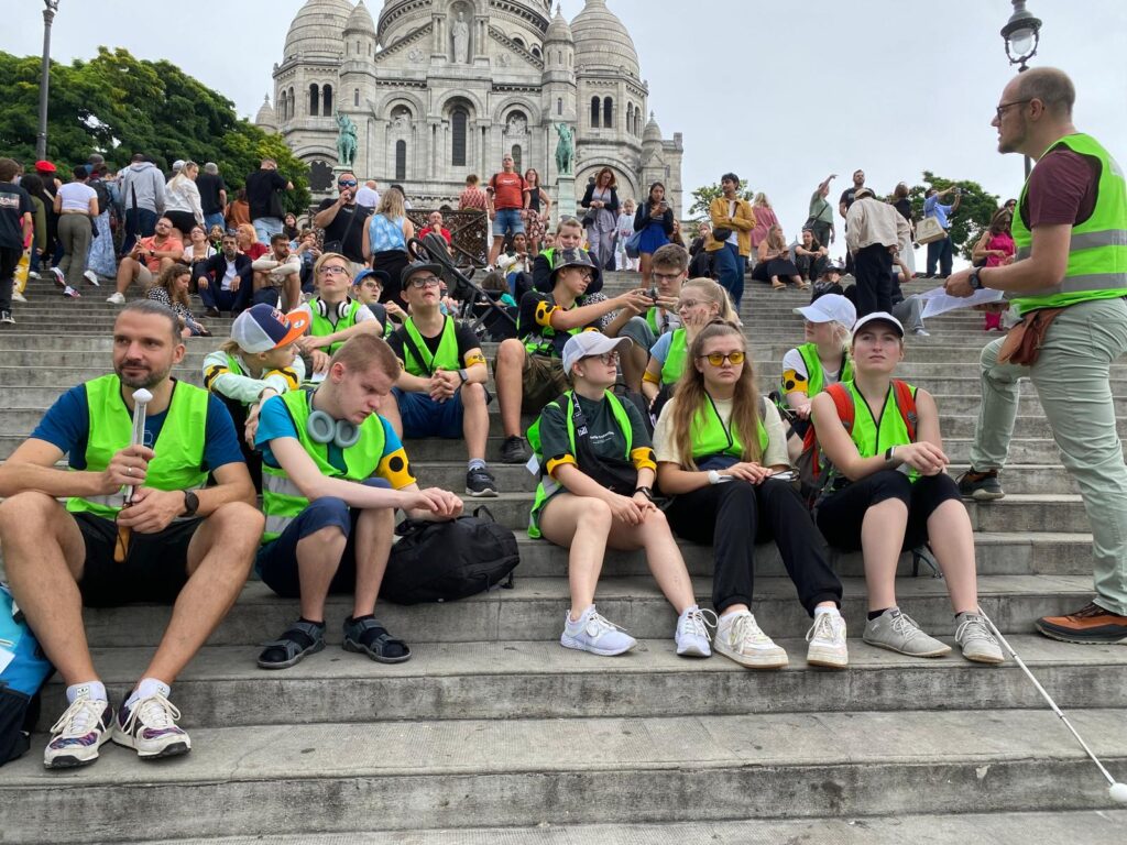 Die Reisegruppe sitzt auf Treppenstufen. Im Hintergrund befindet sich die Basilika Sacre Coeur.