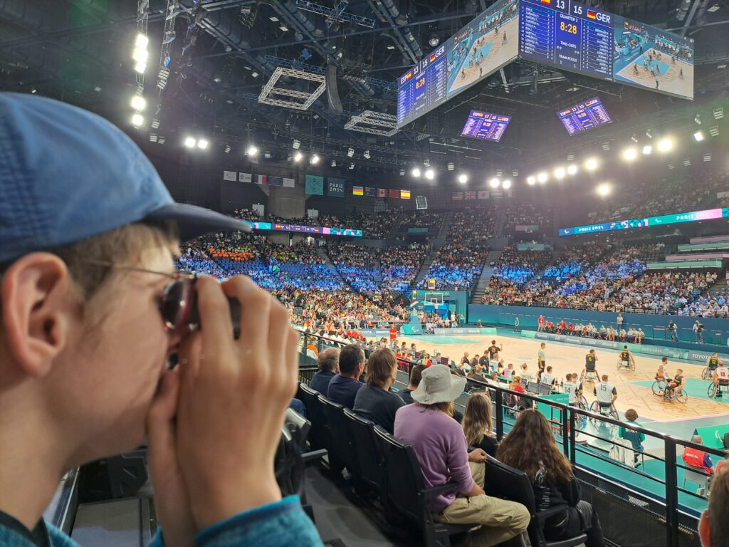 Ein Schüler sitzt auf der Tribüne im Basketballstadion und sieht mit seinem Monokular dem Geschehen auf dem Spielfeld zu. Zwei Rollstuhlbasketballmannschaften bestreiten gerade ein Spiel. Das Stadion ist der gut besucht.