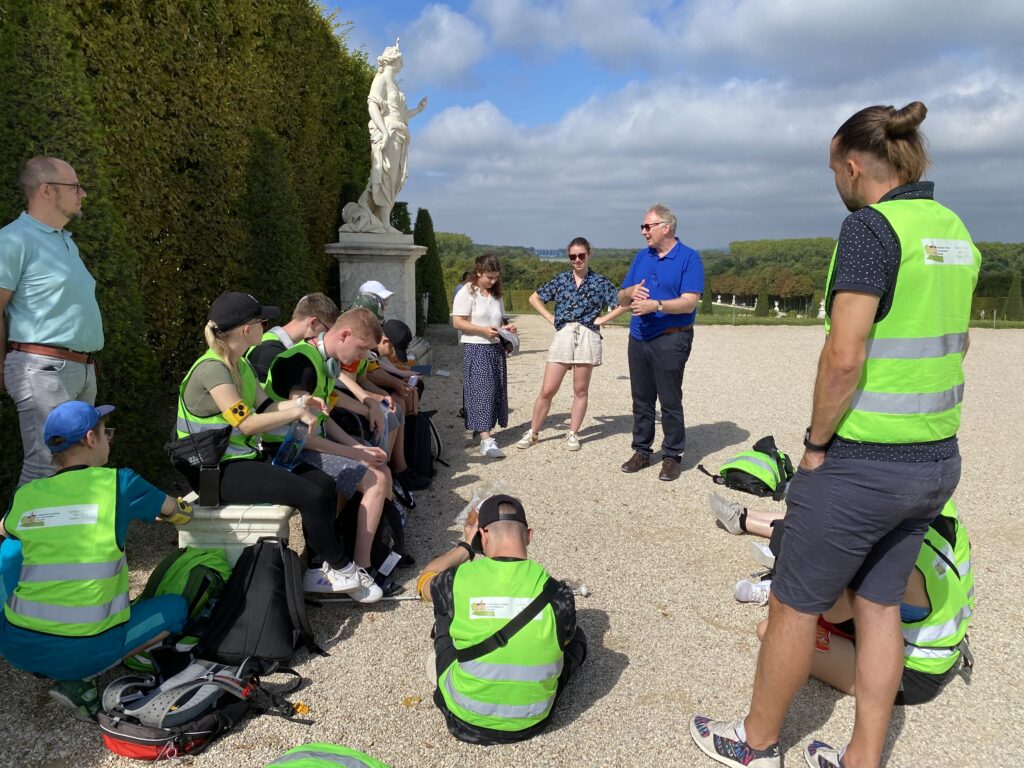 Die Reisegruppe steht im Garten von Versaille und lauscht spannenden Erzählungen.