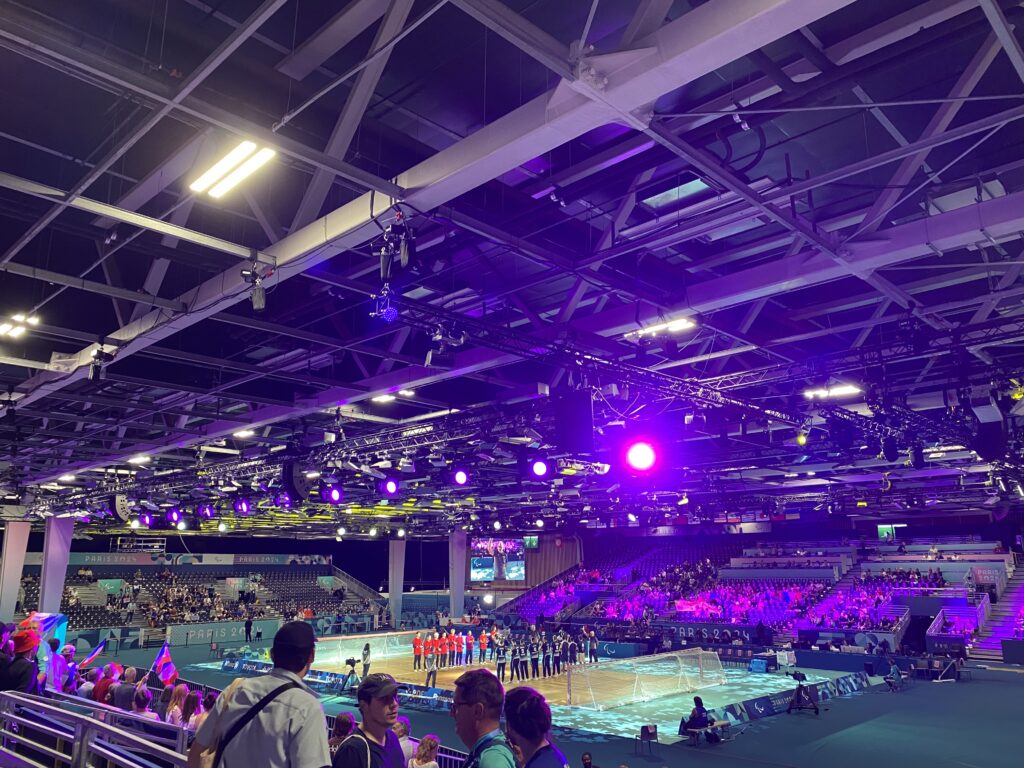 Der Blick in eine Sporthalle, in der Goalball gespielt wird. Beide Mannschaften stehen sich auf dem Spielfeld zur Eröffnung gegenüber. Auf den Rängen sitzen einige Zuschauer.