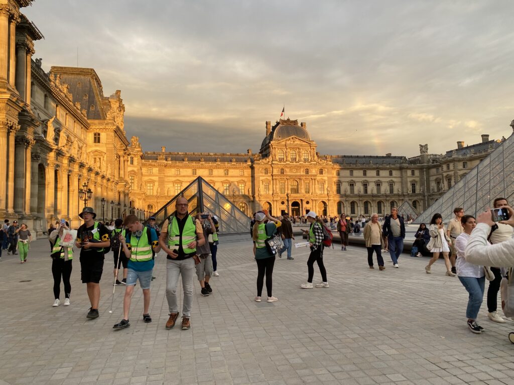 Die Reisteilnehmer in ihren grünen Westen laufen über einen Platz, im Hintergrund der Louvre inklusive der Glaspyramide