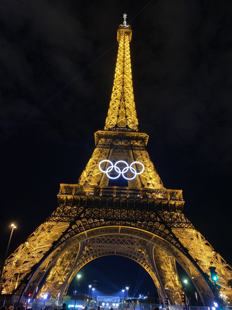 Der beleuchtete Eiffelturm bei Nacht, zusätzlich leuchten die olympischen Ringe.